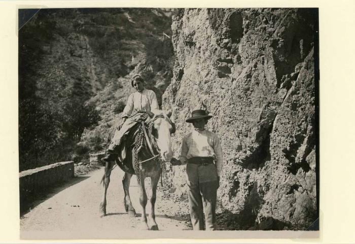 [Homme guidant une mule bâtée sur laquelle est assise une jeune femme, sur la route de Péone, 2e quart du 20e siècle.]